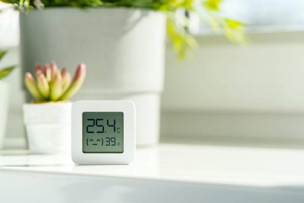 Digital thermometer and hygrometer showing 25.4°C and 39% humidity on a white surface with plants in the background.