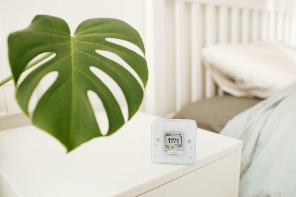 Digital CO2 monitor showing 1171 ppm on a white bedside table next to a large green monstera leaf.